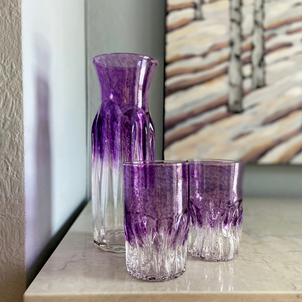 Carafe and two glasses with clear bottom and purple glass top on a white marble surface with a picture in the background.