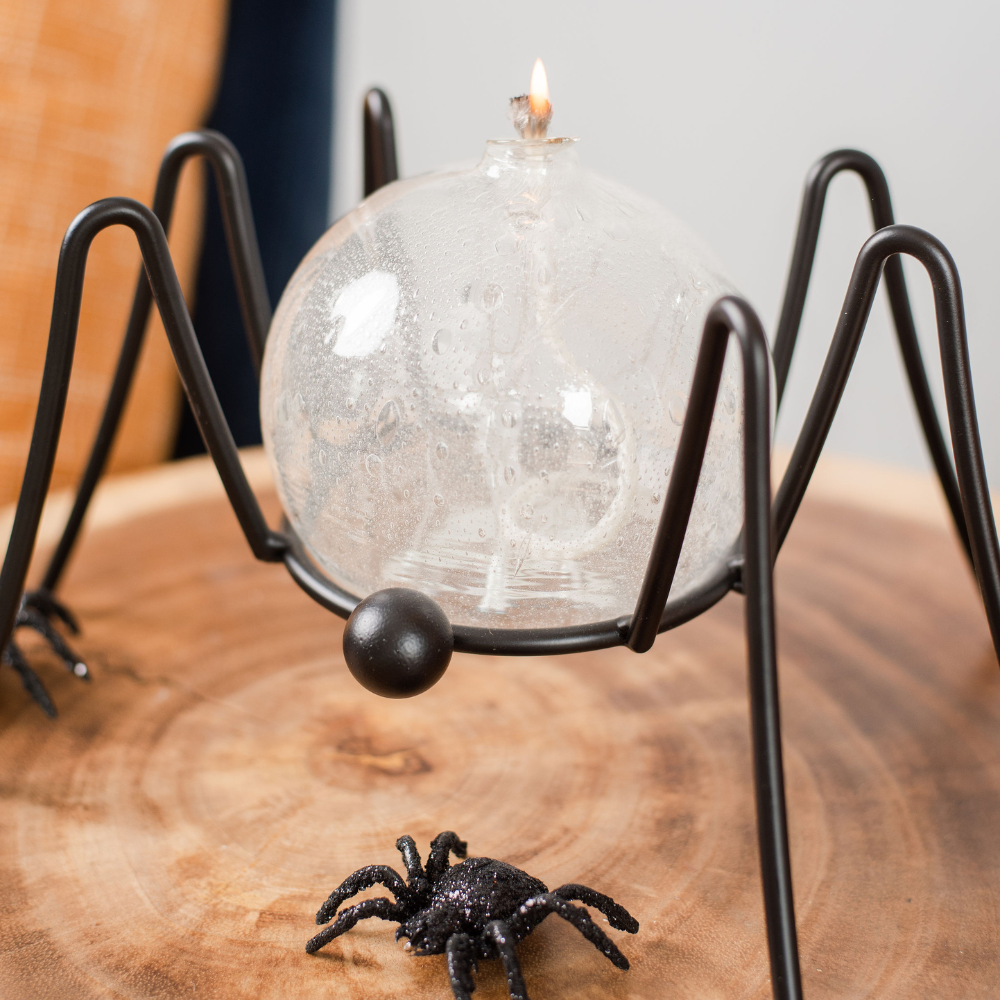 Lit clear glass orb with a wick and glass webbing pulled from bottom to displayed in a black spider garden holder on a wooden table.