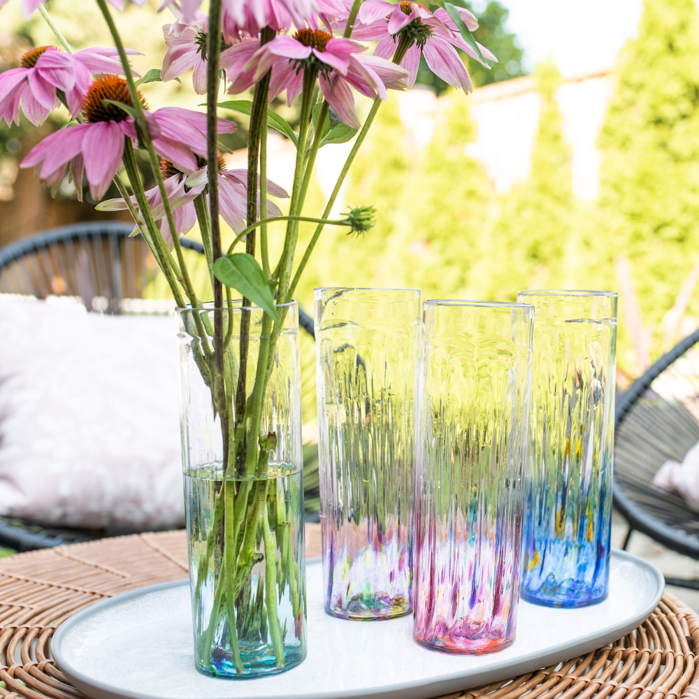 Four clear glass vases, with teal, purple, pink and blue on the bottom and multicoloured dots throughout. Pink flowers with green stems in teal vase. All on a white tray with greenery and patio chairs in the background