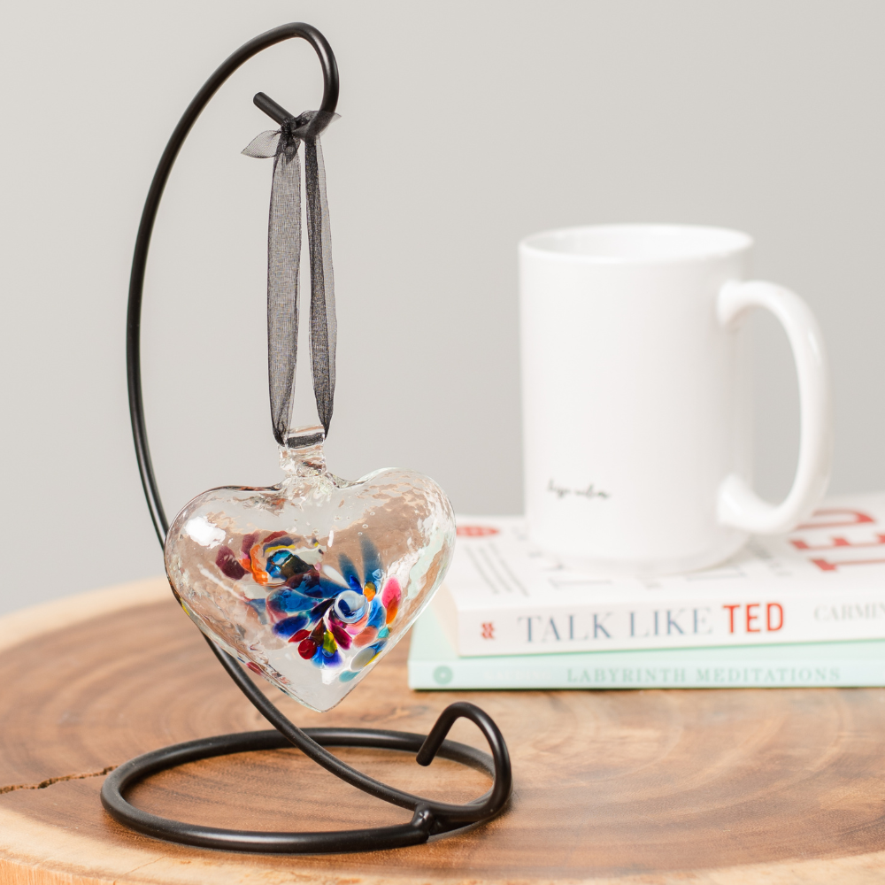 Clear glass heart with blue, yellow, purple, pink, orange and opaque white glass swirled in the centre hanging from a black ribbon on a black curved single ornament holder. Displayed on a wood table with a white book and mug to the right. 