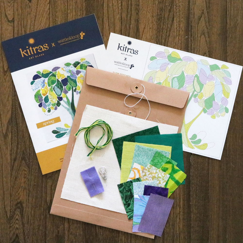 Green, purple and blue fabrics and threads on a brown envelope with slow stitch pattern in background on a wood floor