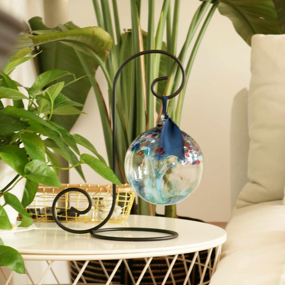 Clear glass orb with blue glass pulled through the middle and yellow, red, purple, pink, orange, green and blue bits on top on a black ornament stand. Displayed on an end table surrounded by greenery. 