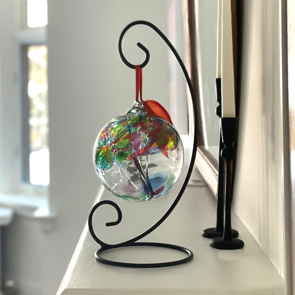 Clear glass orb with green glass pulled through the middle and red, green, lime, blue and pink glass bits on top on a black curved ornament stand displayed on a white mantel. 